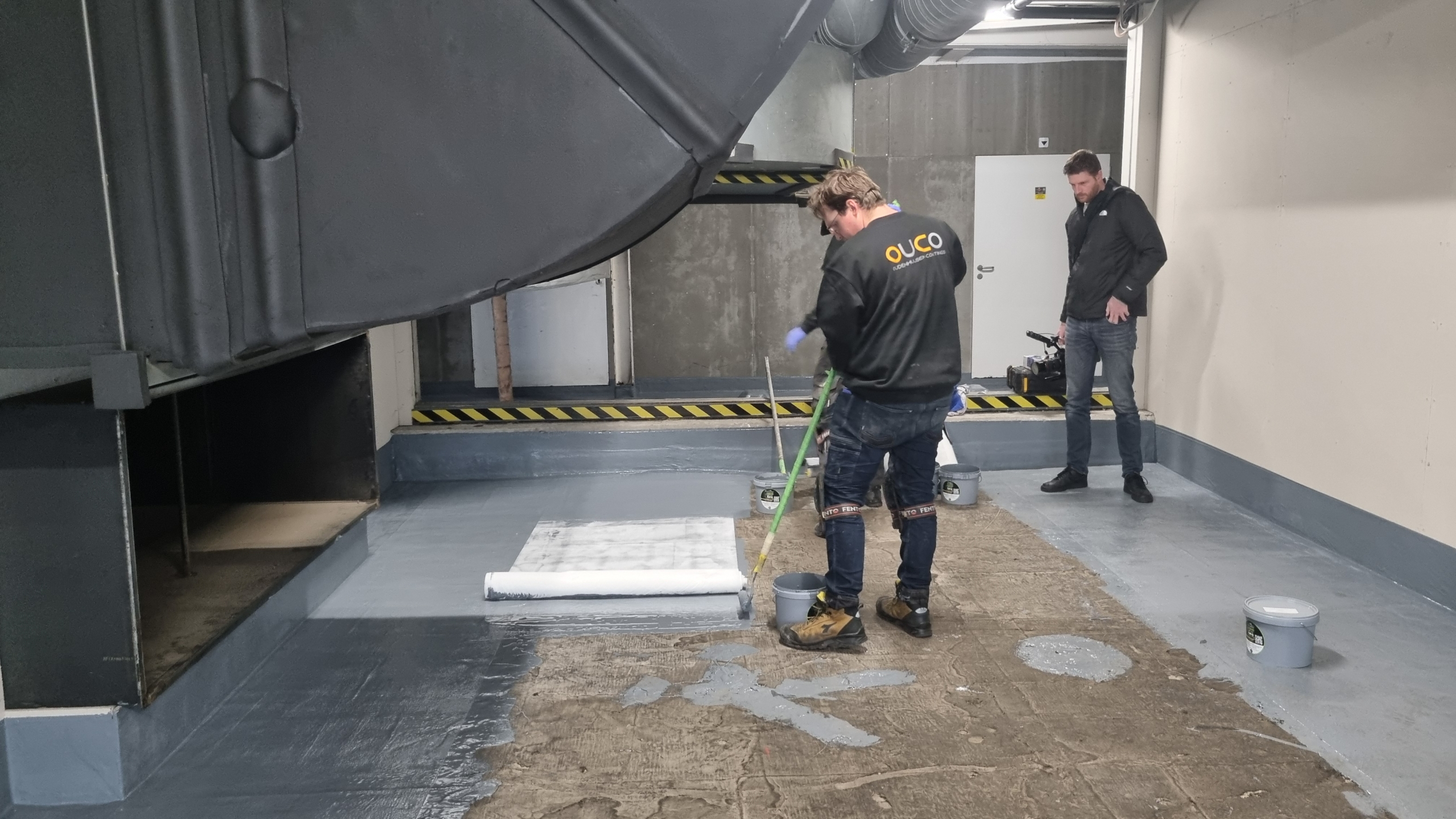 Applicateur rolt vloeibare afdichting met wapeningsvlies op een betonvloer, terwijl een collega meekijkt.