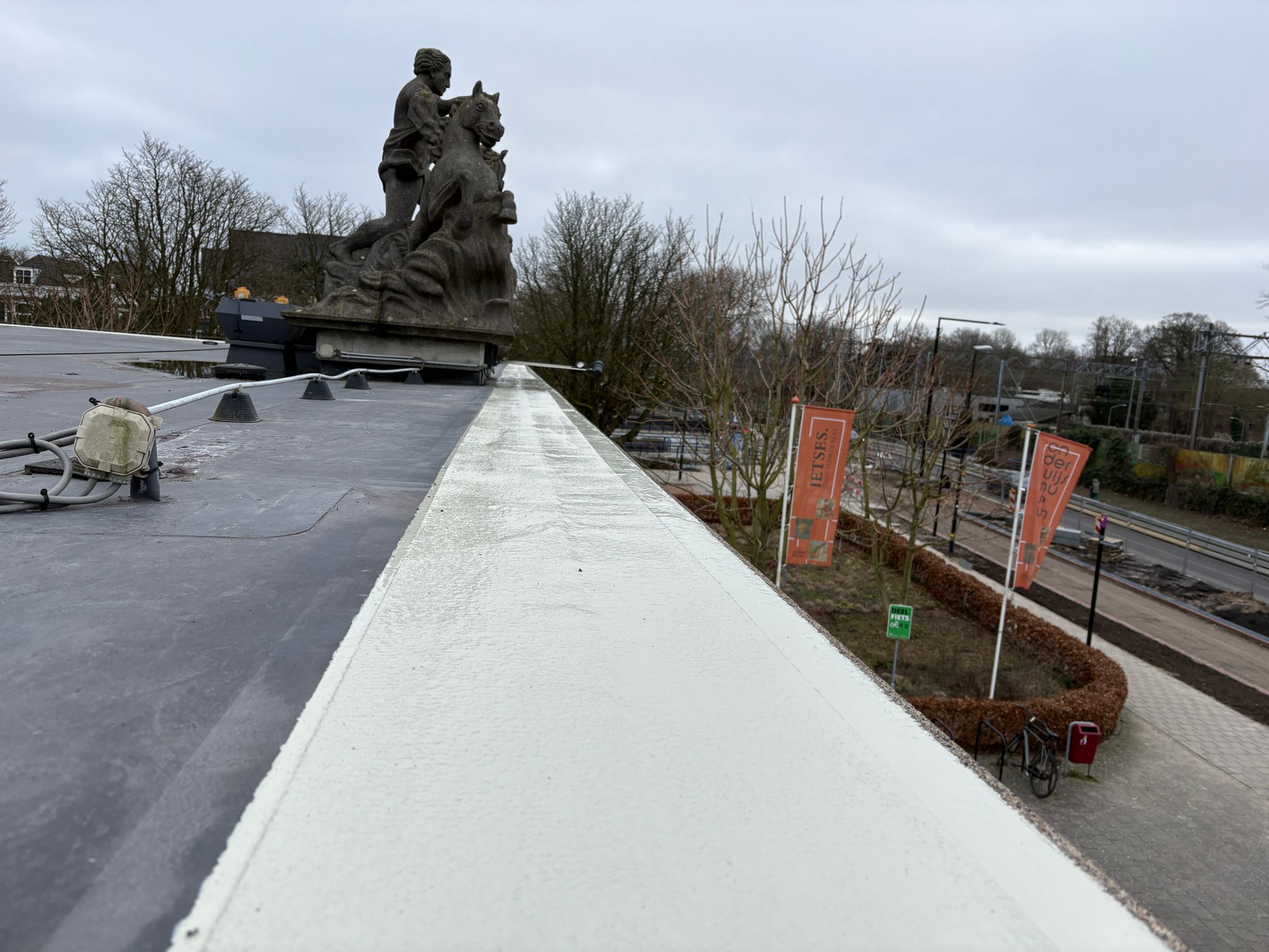 Dakrand in wit afgewerkt met vloeibare dakbedekking naast een plat dak met beeldengroep op de hoek.
